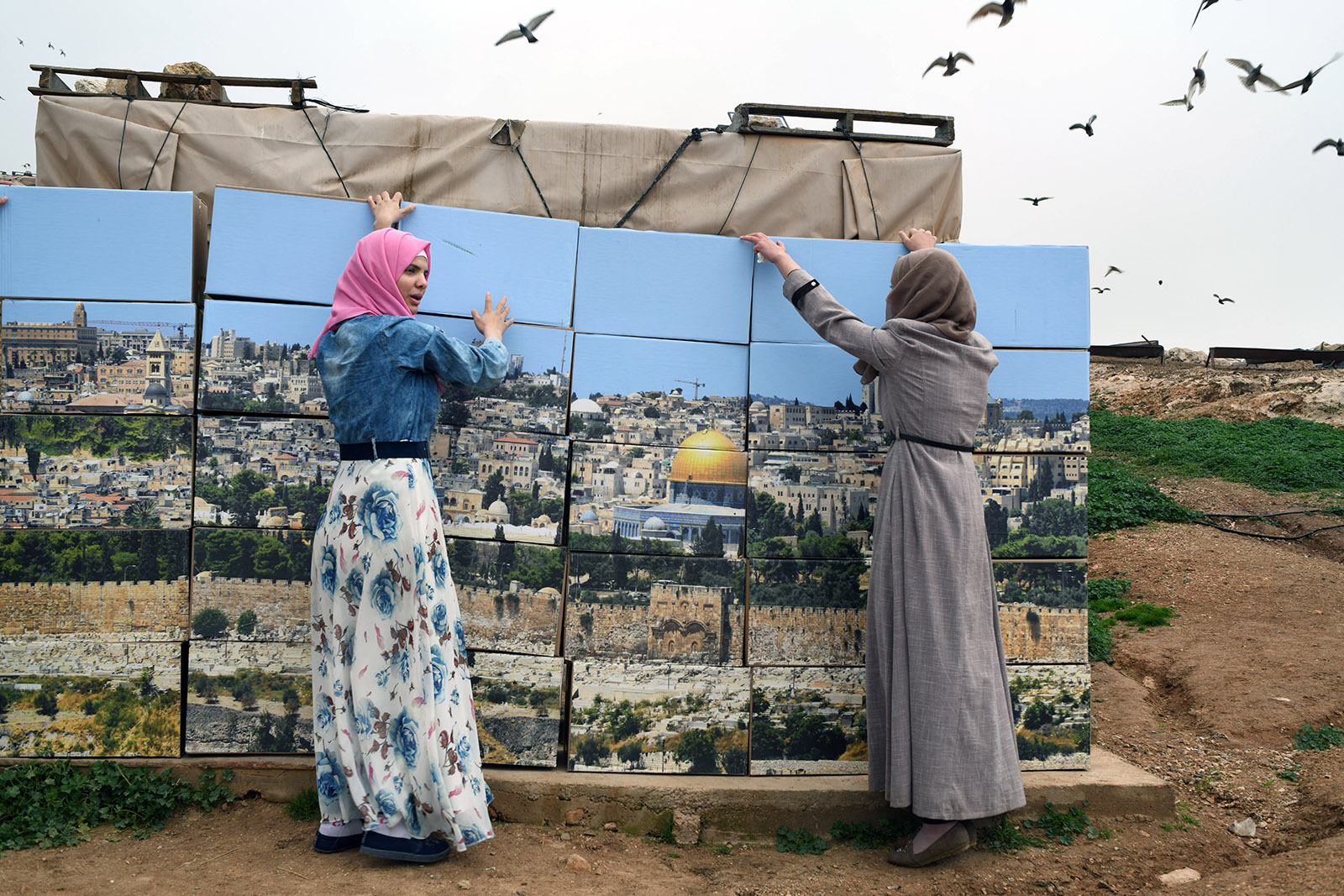 Two Palestinian women stand beside a patchwork image of the Al-Aqsa Mosque in Jerusalem. The photograph is printed at body-height scale, and split into circa 30 centimetre-wide segments. Around the women, birds fly through a grey sky